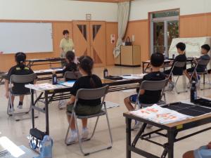 書道講座開催の挨拶をする先生と座って話を聞く参加者